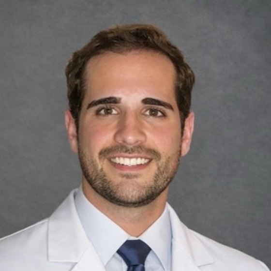 A man with short dark hair and a trimmed beard, in a white coat and dark tie.